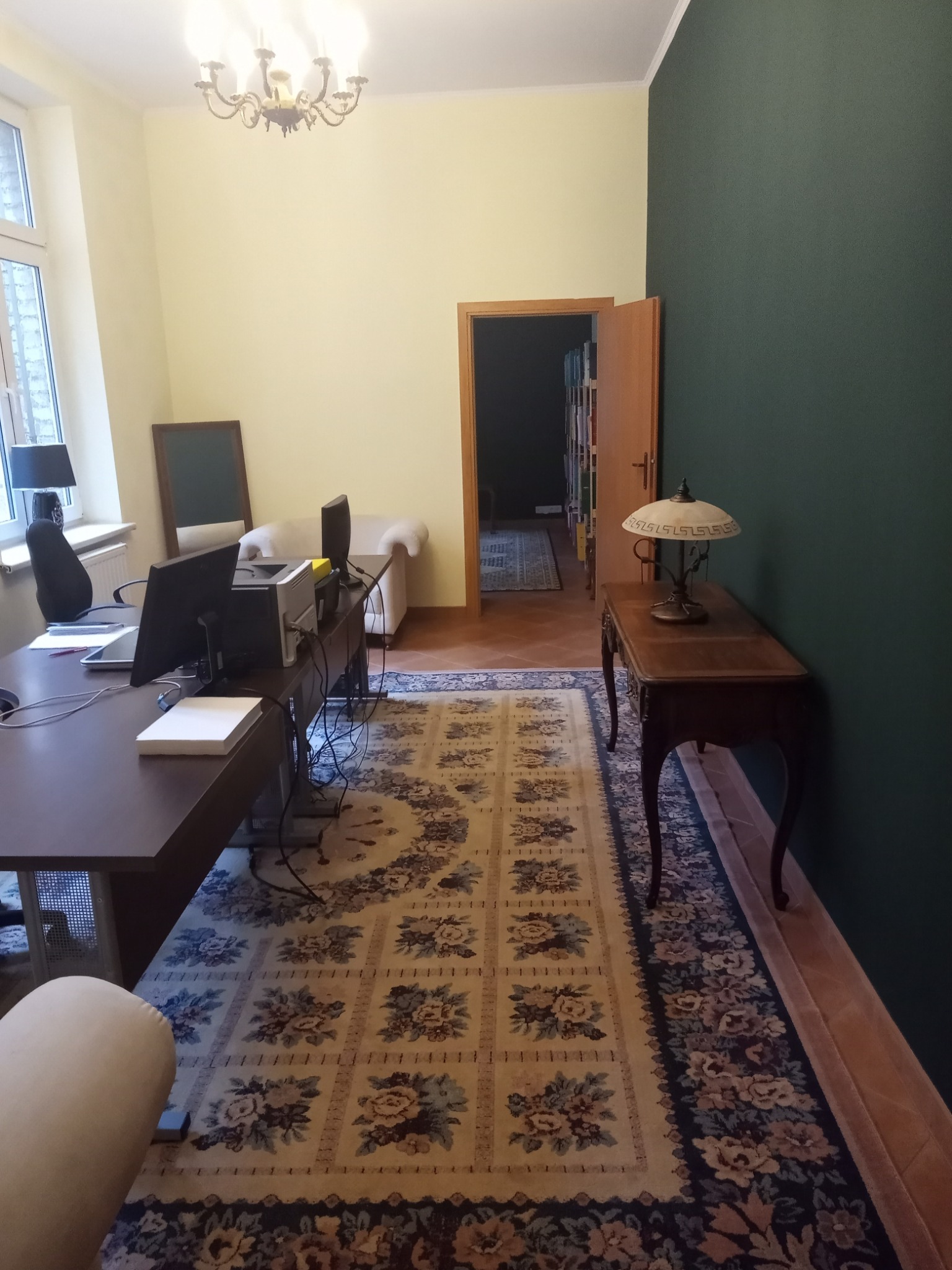 Wnętrze biura z ciemnym biurkiem, komputerami, kremowym dywanem w kwiaty i antycznym stolikiem z lampką, widok na pokój z biblioteczką.