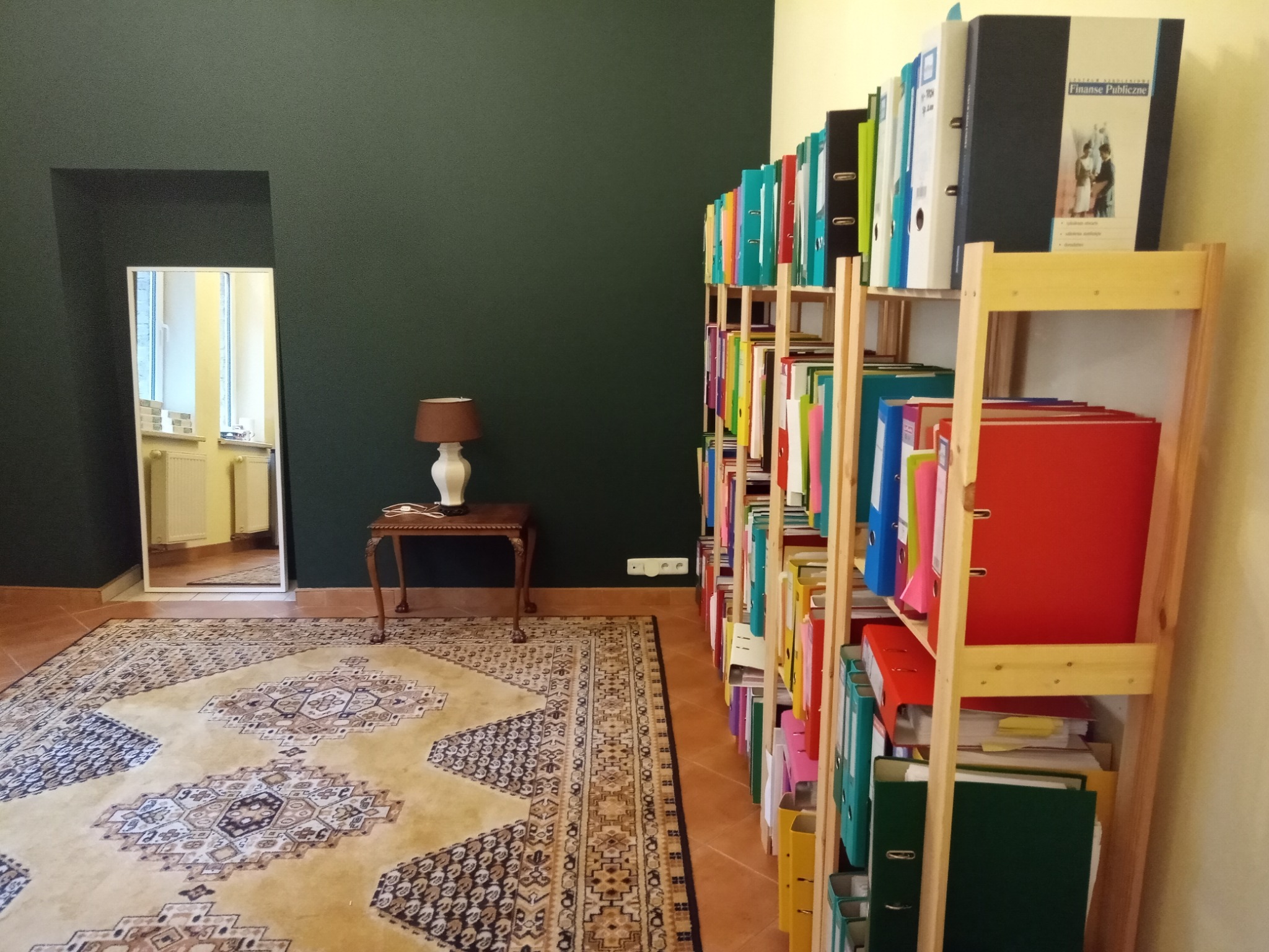 Regał wypełniony kolorowymi segregatorami z dokumentami, ustawiony w eleganckim wnętrzu z ciemnozieloną ścianą i dywanem w orientalne wzory. W tle widoczne lustro odbijające fragment pomieszczenia.