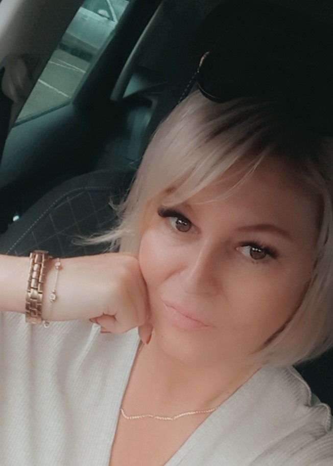 Portret kobiety z blond włosami i delikatnym makijażem, siedzącej w samochodzie w Katowicach, z widocznym złotym zegarkiem i łańcuszkiem na szyi.