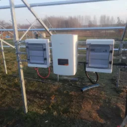 Instalacja paneli słonecznych: widok na metalową konstrukcję, biały inwerter Solis i dwie skrzynki elektryczne z przezroczystymi drzwiczkami na zewnątrz, na tle pola i drzew.