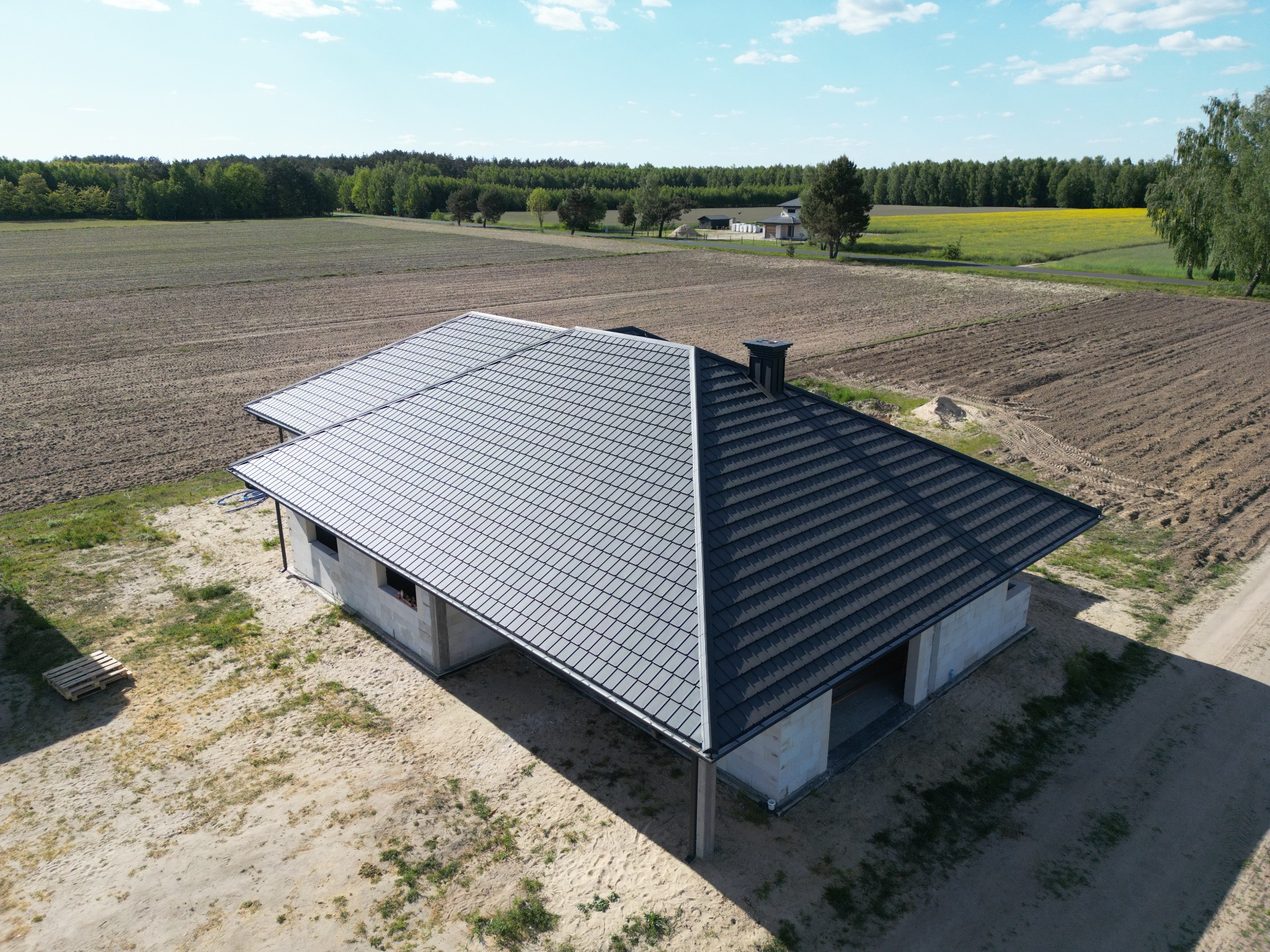 Nowy, grafitowy dach z dachówki ceramicznej na surowym domu jednorodzinnym w Łomazach, widok z lotu ptaka na tle pól i lasów.