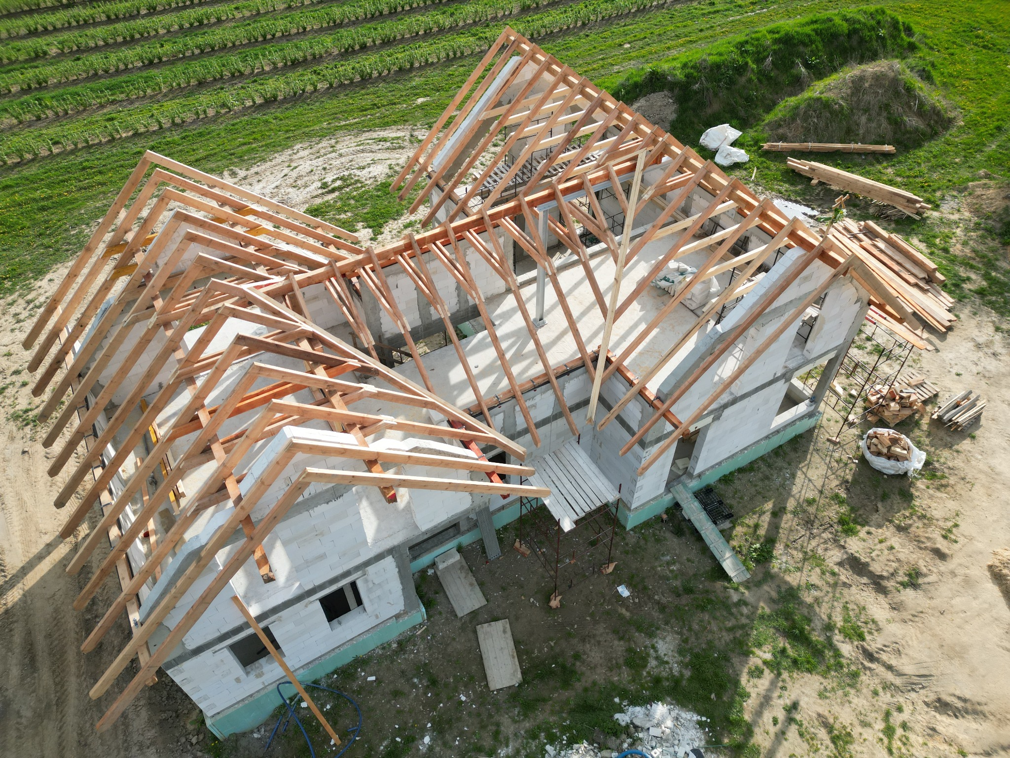Widok z lotu ptaka na budowę domu jednorodzinnego z częściowo ukończoną drewnianą konstrukcją dachu na tle zielonego pola uprawnego.