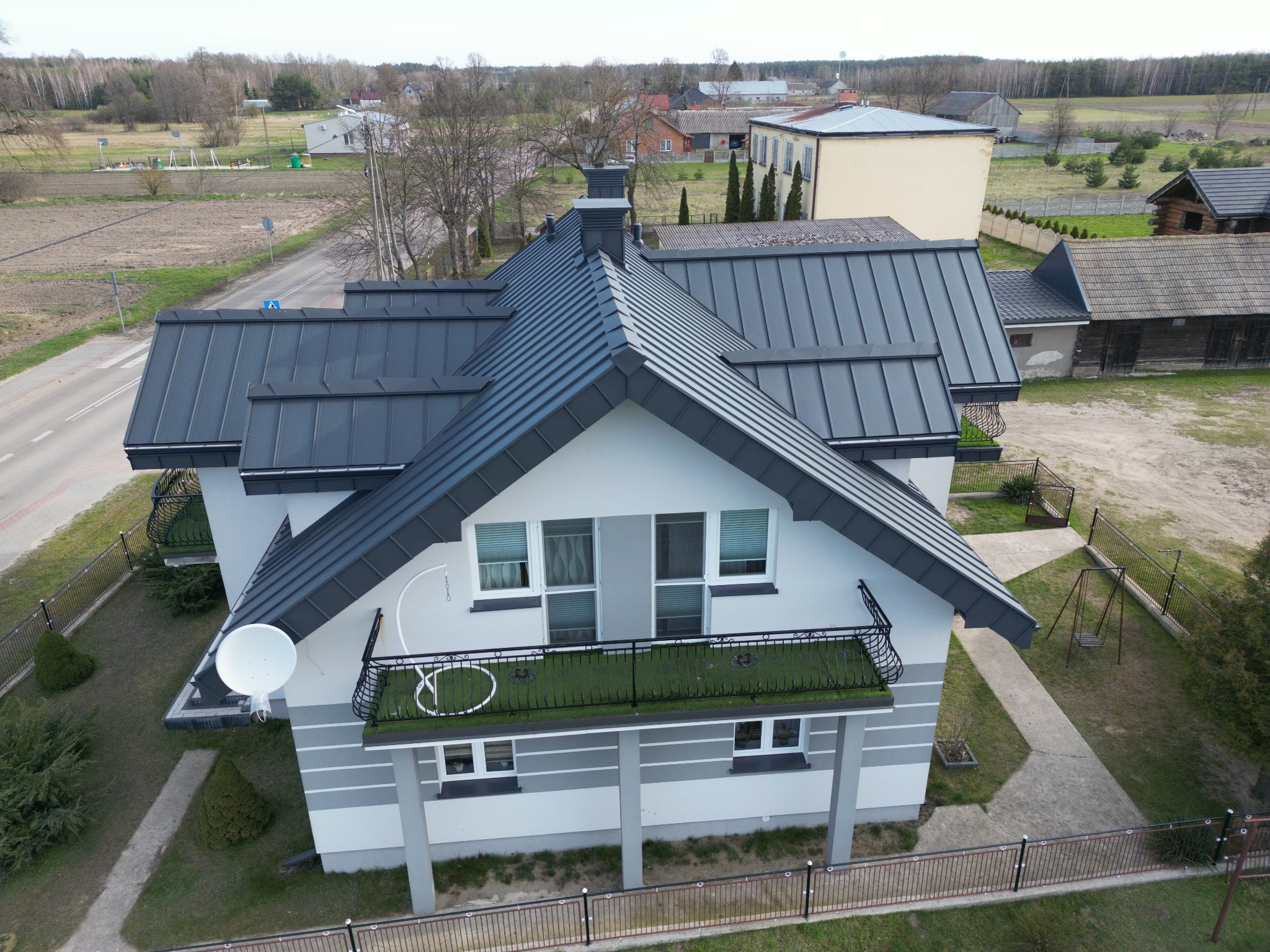 Dom jednorodzinny z ciemnoszarą, blachodachówką, widok z lotu ptaka, w tle zabudowa wiejska i droga.