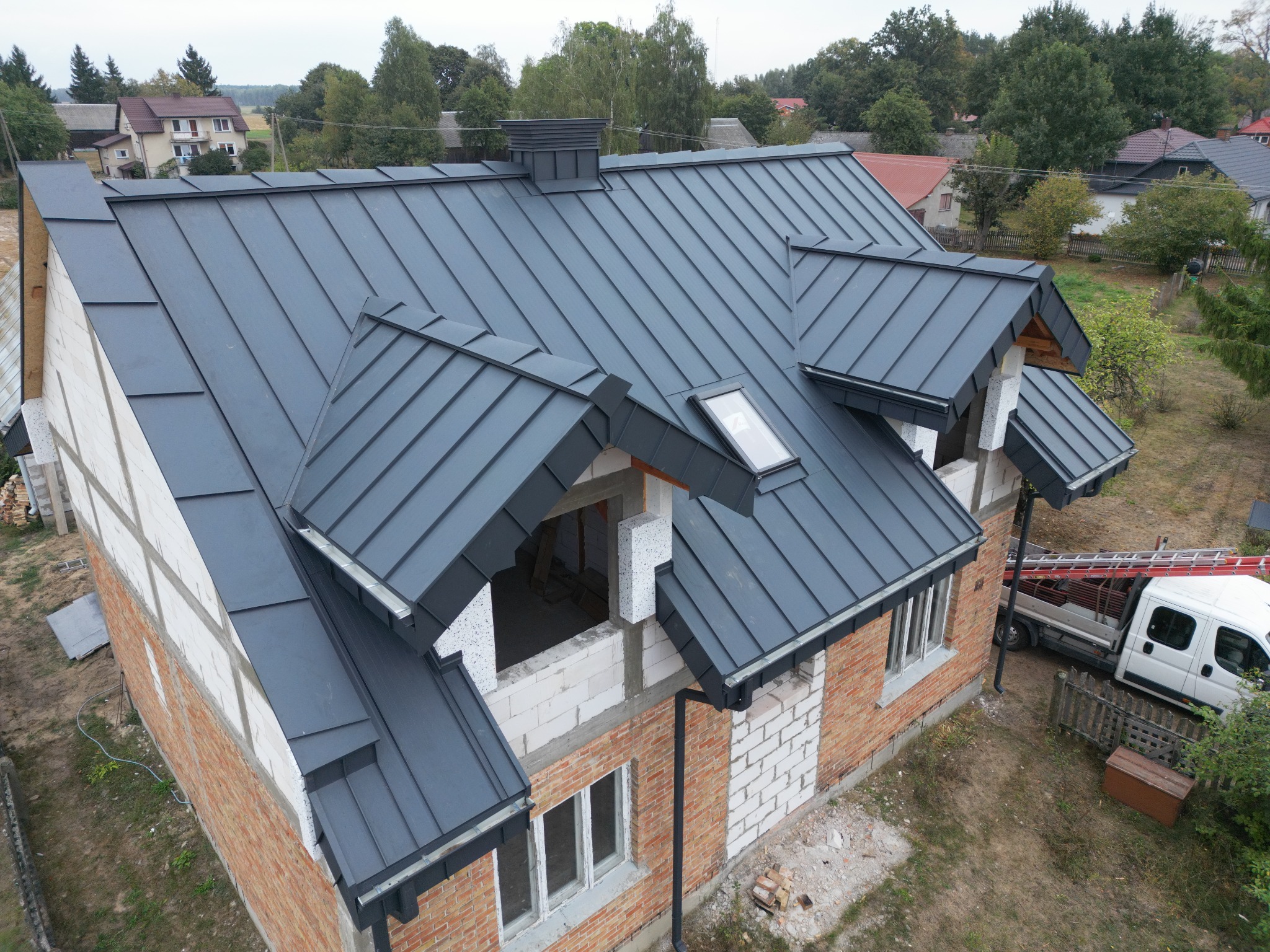 Nowy, grafitowy dach z blachy na domu w trakcie budowy, widoczne okna dachowe, rynny i częściowo wykończona elewacja z cegły i bloczków.