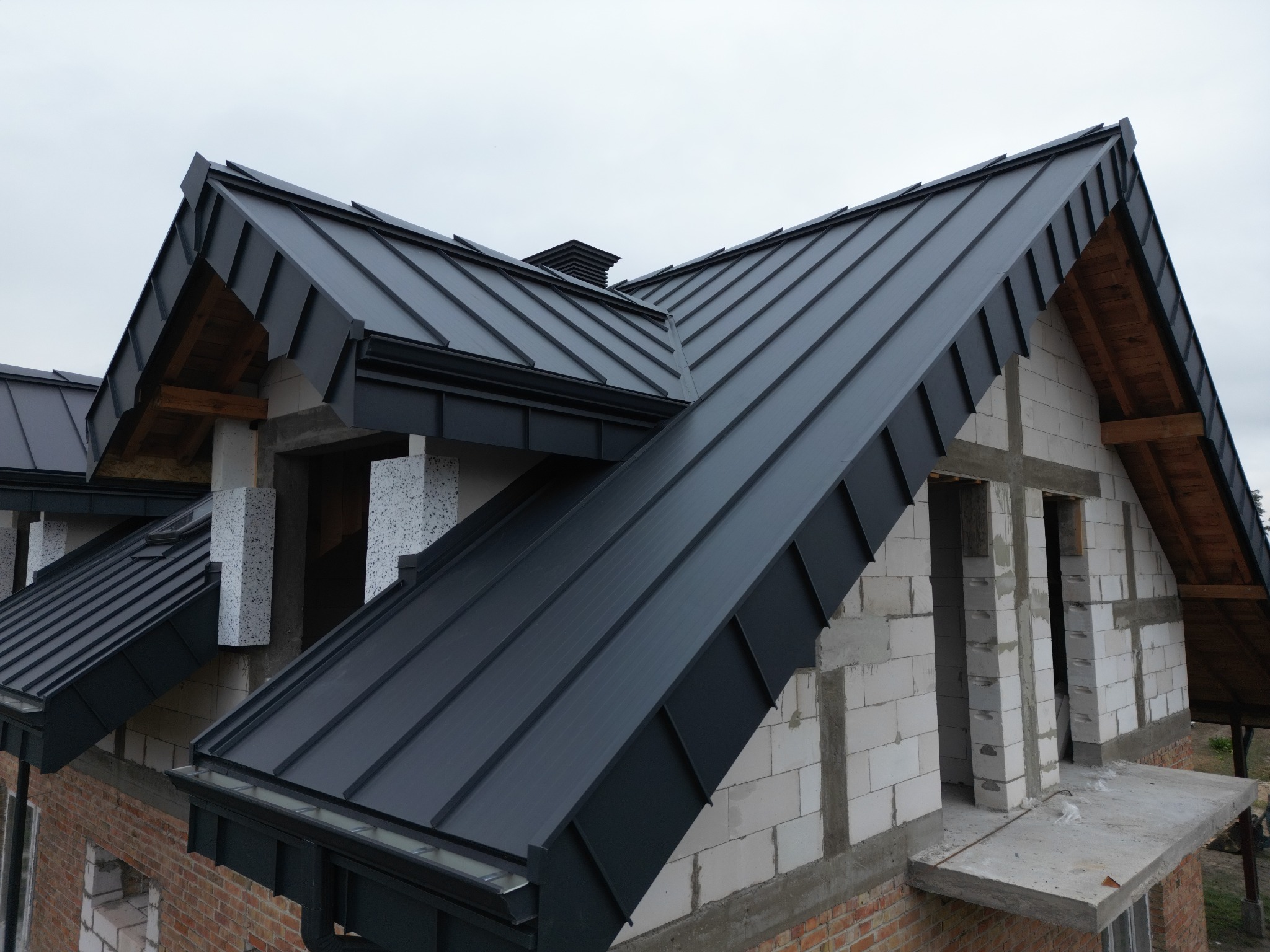 Nowoczesny, ciemnoszary dach z blachy na budynku w trakcie budowy, widoczne białe bloczki i czerwona cegła, elementy drewniane więźby i komin.