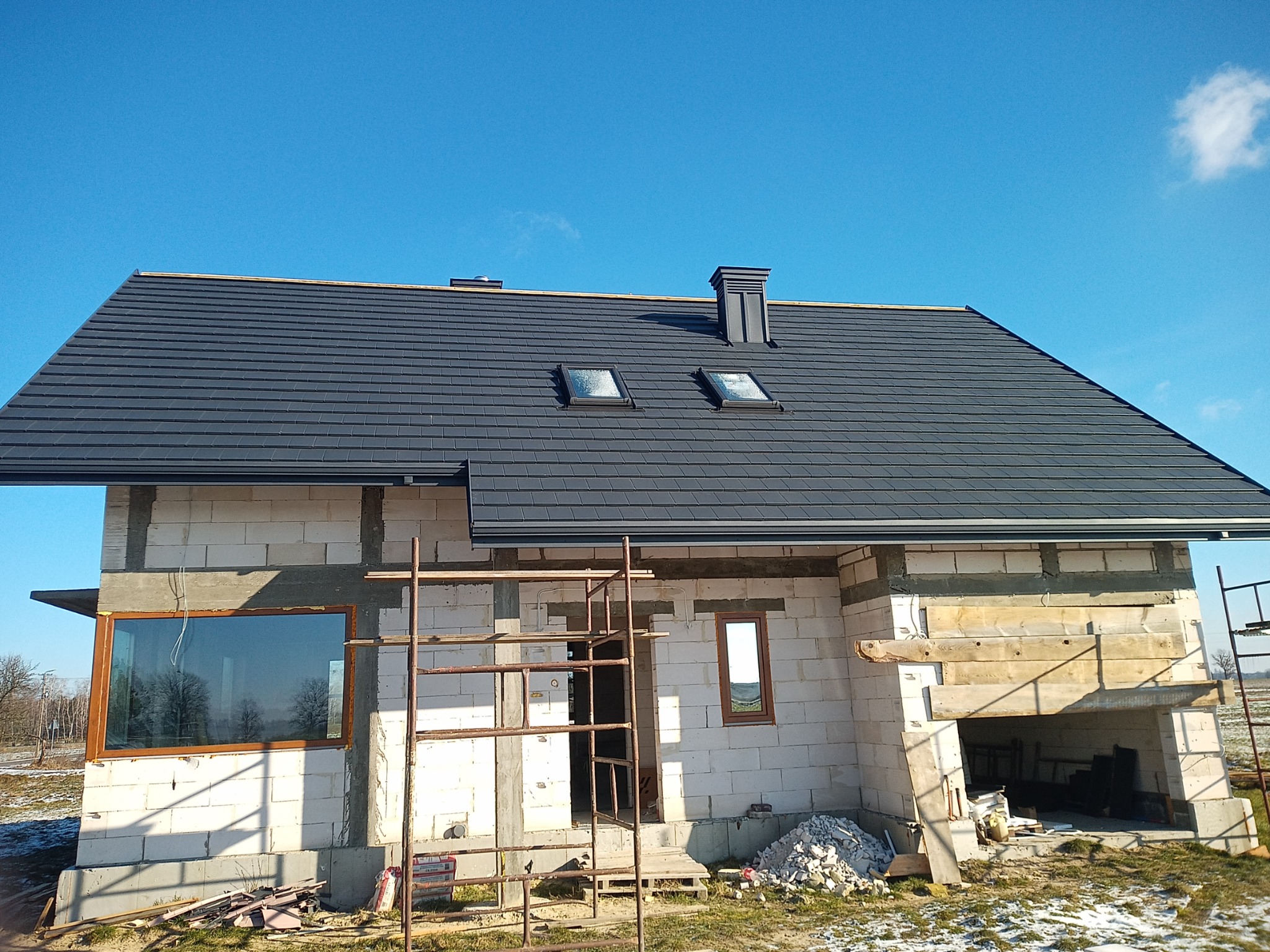 Dom w trakcie budowy z zamontowanym, grafitowym dachem z dwoma oknami dachowymi i kominem, widoczne rusztowania i teren budowy.