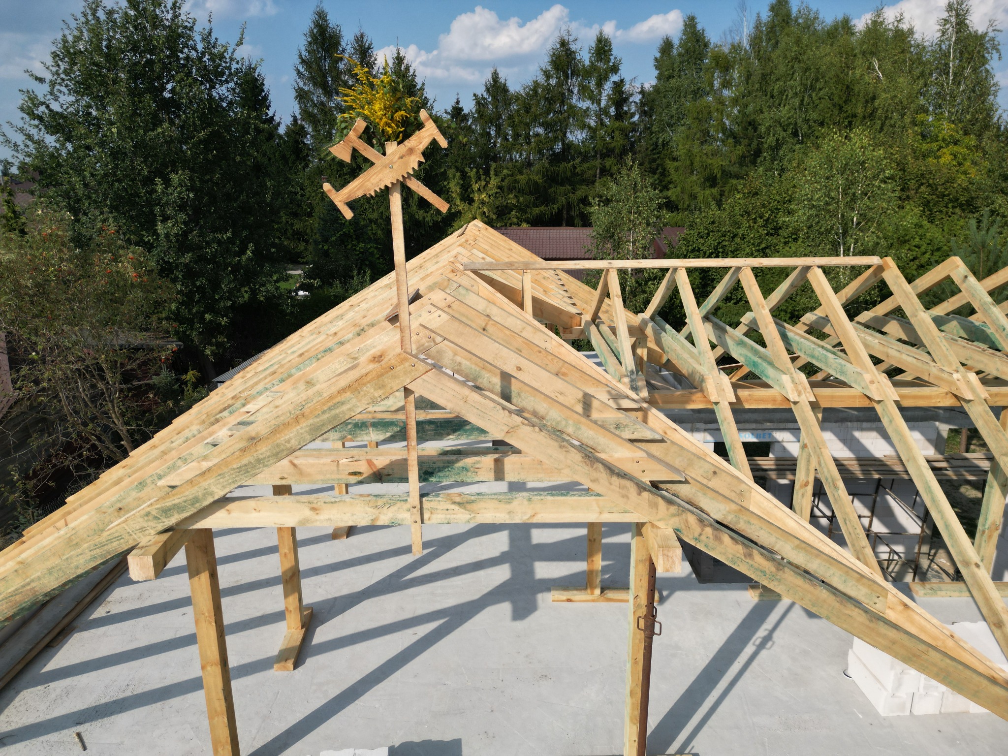 Wznoszenie drewnianej konstrukcji dachu z dekoracyjnym symbolem cieśli na szczycie, widok z góry, w tle zielone drzewa i błękitne niebo.