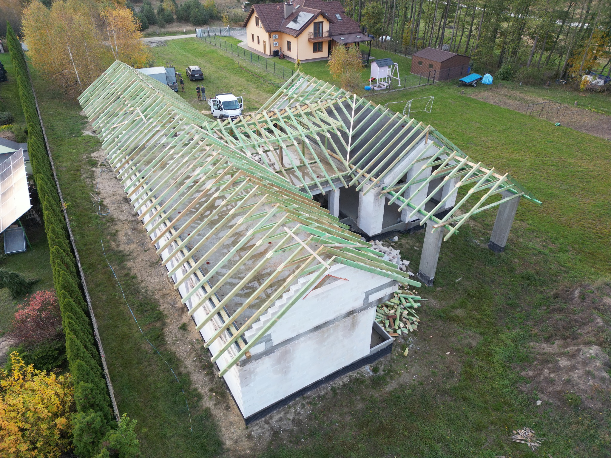 Widok z drona na budowę domu jednorodzinnego z widoczną konstrukcją drewnianą dachu w trakcie montażu, na działce leżą sterty przyciętego drewna, w tle zaparkowany biały bus ekipy budowlanej...