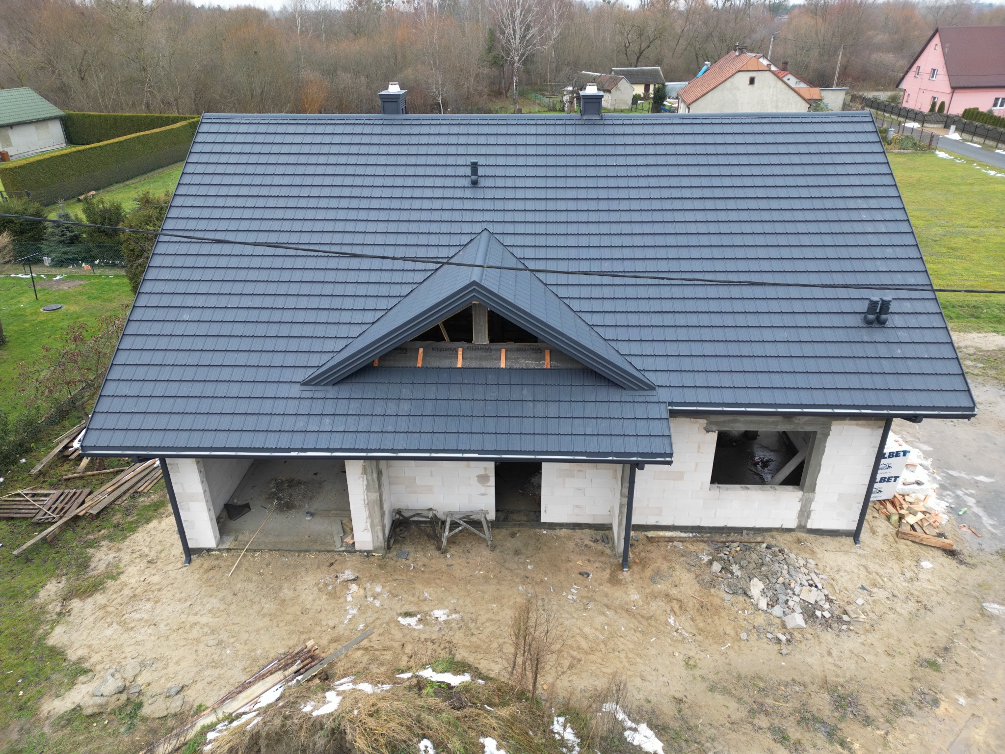 Widok z góry na dom w trakcie budowy z nowym, ciemnoszarym dachem z dachówki, widoczna więźba dachowa na szczycie, surowe ściany z betonu komórkowego i teren wokół domu pokryty piaskiem i resztkami...