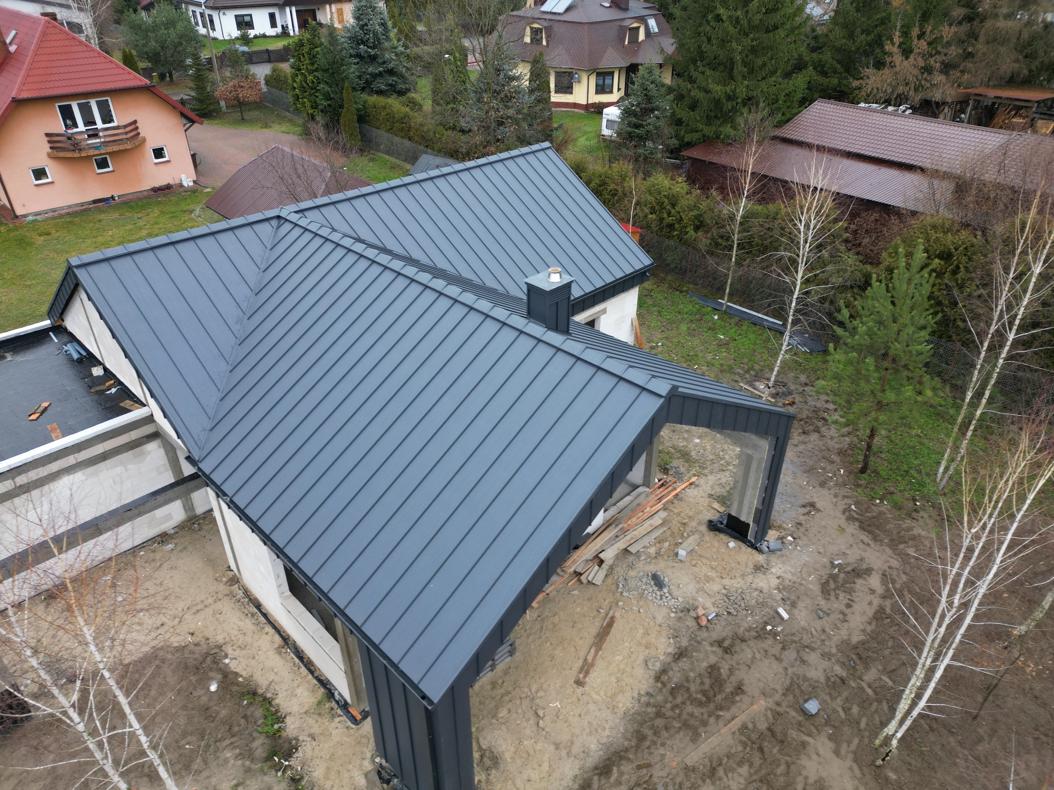 Nowoczesny dom w trakcie budowy z ciemnoszarą blachą na dachu, widok z góry na ukończony dach i komin, surowy stan ścian, teren wokół w trakcie prac ziemnych.