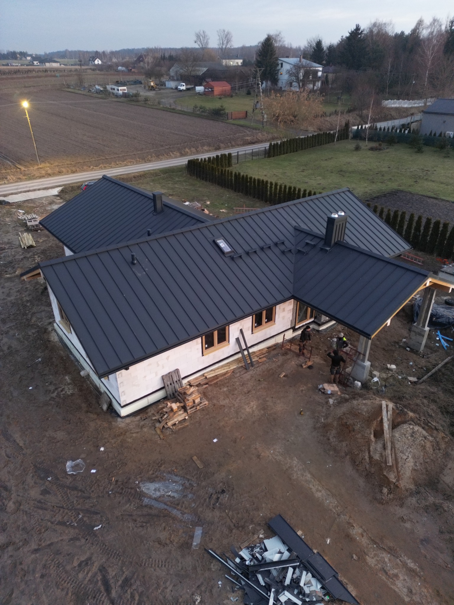 Widok z góry na dom w trakcie budowy z ciemnoszarą blachodachówką, widoczne okna z drewnianymi ramami, komin, ziemia wokół domu oraz trzech pracowników przy konstrukcji zadaszenia tarasu.