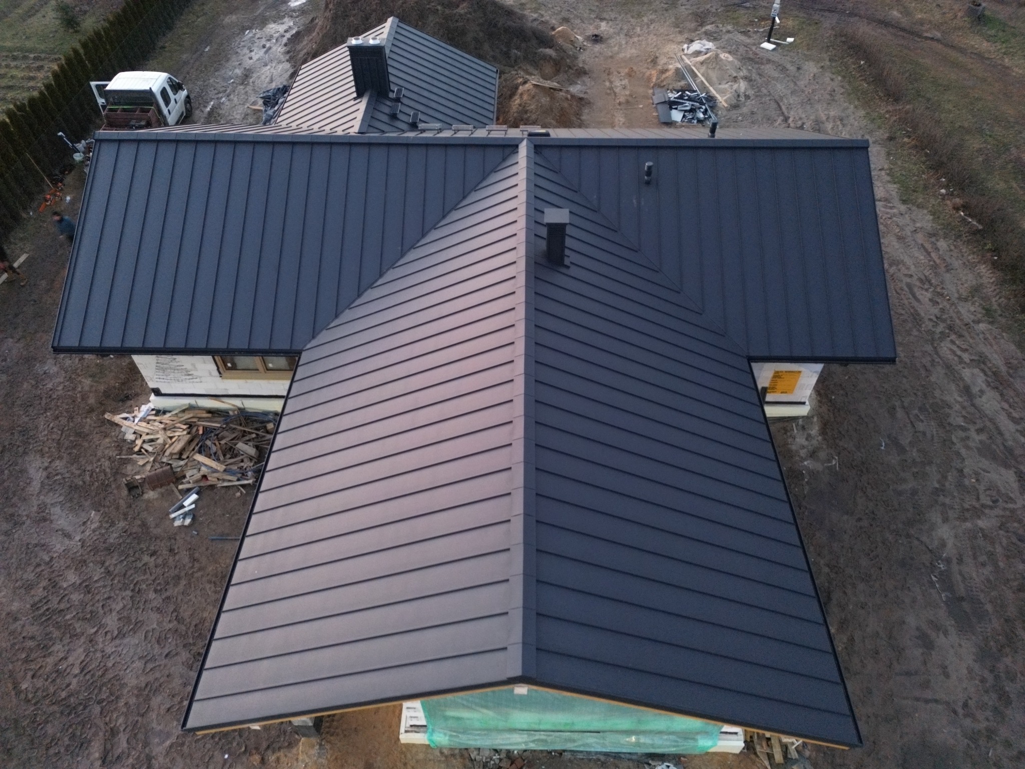 Nowoczesny dom z ciemnoszarym, metalowym dachem panelowym, widok z góry na ukończony dach z kominami, teren budowy wokół.