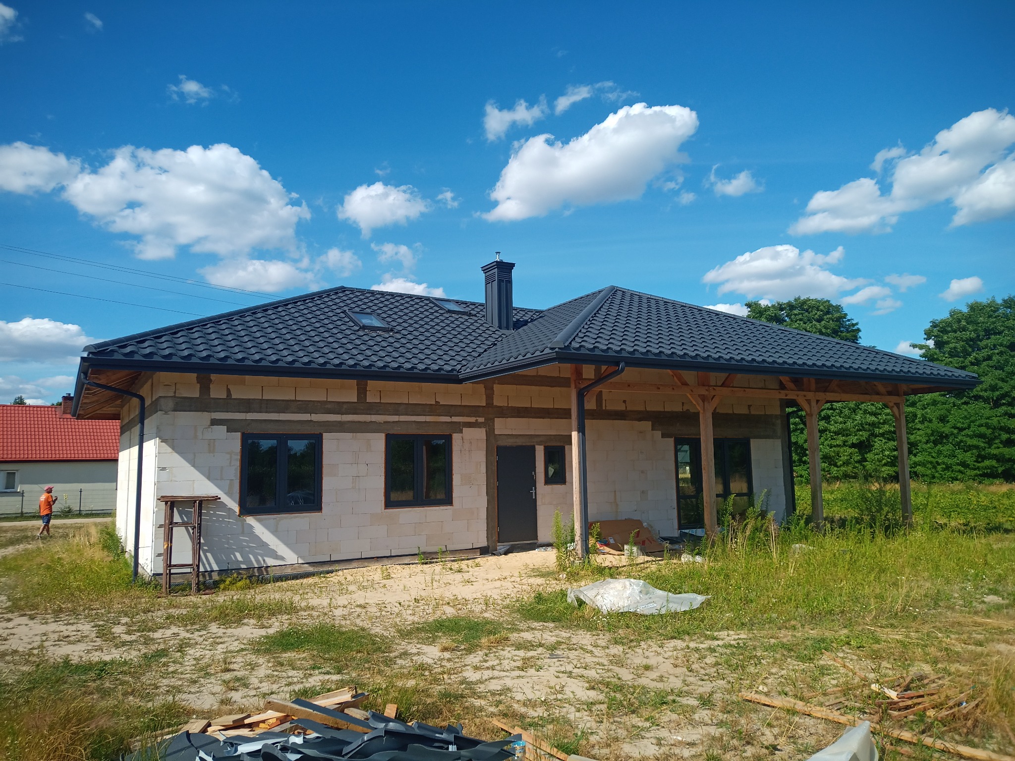 Niedokończony dom jednorodzinny z czarnym dachem z blachodachówki i ciemną stolarką okienną, widoczne fragmenty rynien, na pierwszym planie składowane elementy blacharskie.