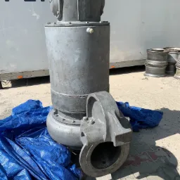 Duży, szary element przemysłowy z odłączoną częścią, leżący na niebieskiej plandece na zewnątrz, prawdopodobnie przed transportem lub obróbką.