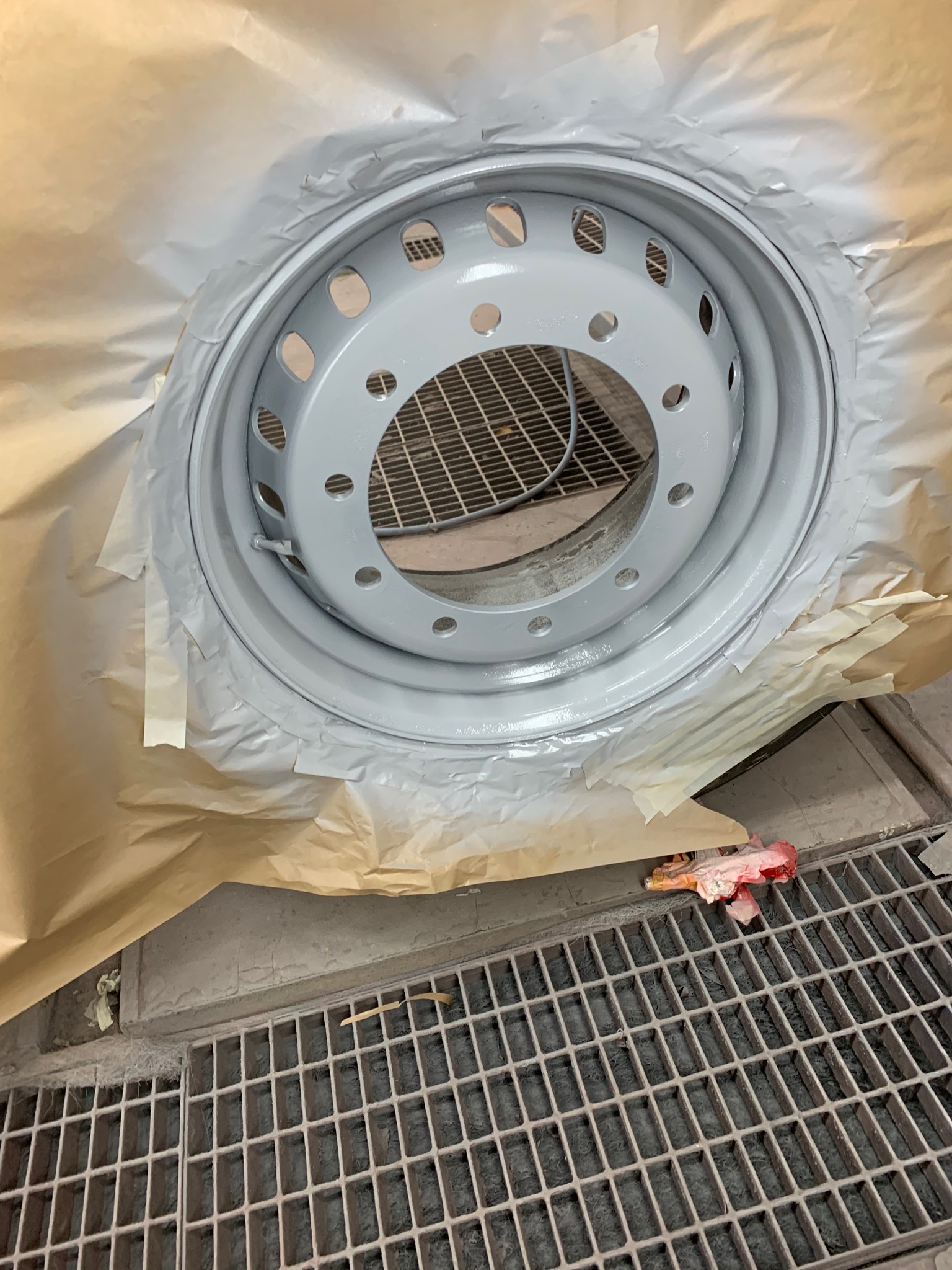 Szara felga samochodowa po świeżym malowaniu, zabezpieczona papierem maskującym, widoczna kratka ściekowa w tle.