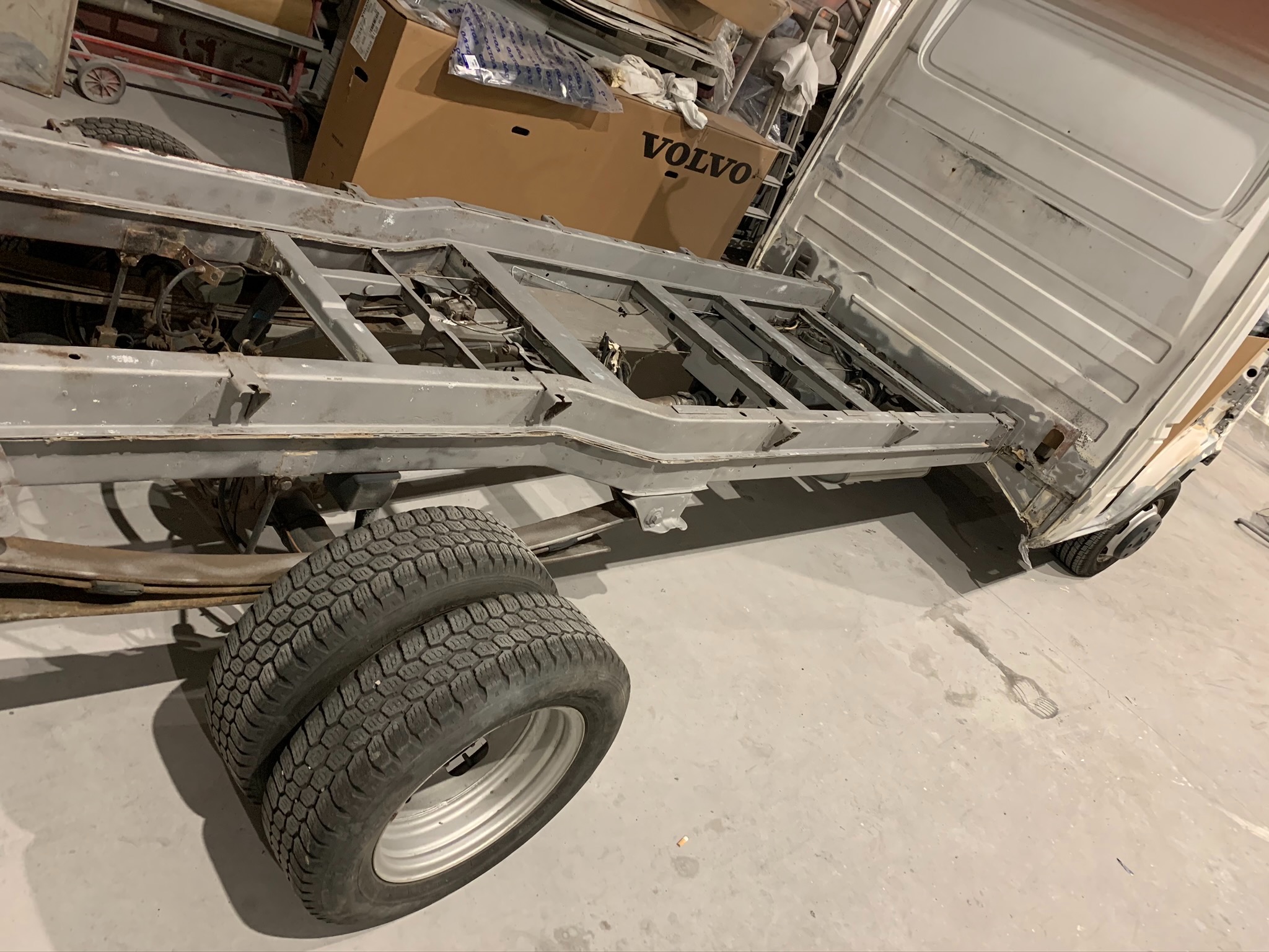 Podwozie samochodu dostawczego przed renowacją, widoczne ślady korozji i starej farby, dwa koła z oponami o agresywnym bieżniku, karton z logo Volvo w tle.