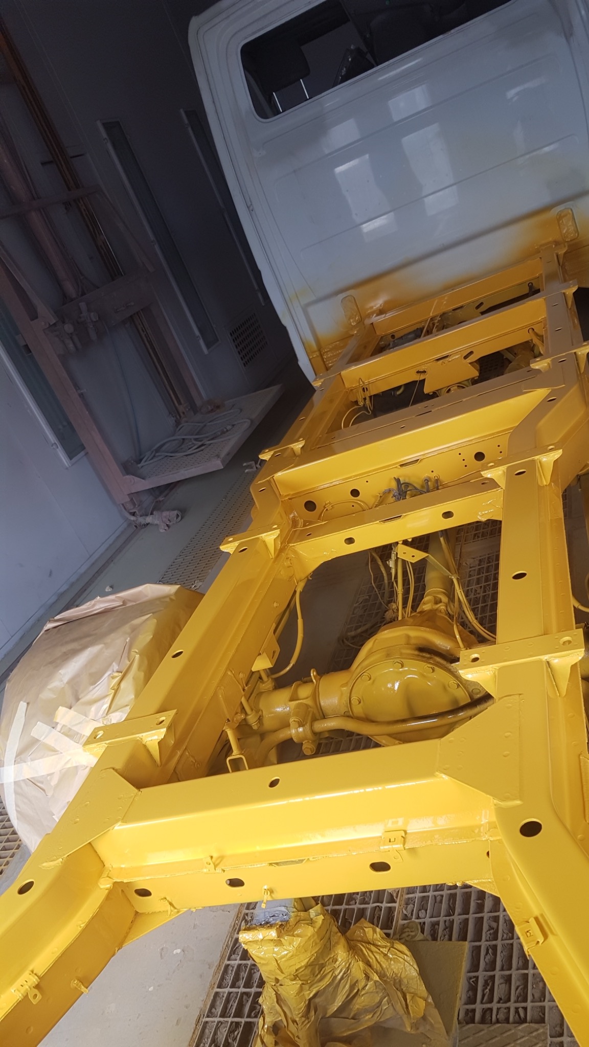 Żółta rama samochodu po nałożeniu farby w kabinie lakierniczej, widoczny fragment karoserii i osłonięte elementy podwozia.