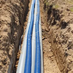 Wykop z ułożonymi szarymi rurami osłonowymi, w których umieszczone są niebieskie rury karbowane, zabezpieczone opaskami zaciskowymi; widok z góry wzdłuż wykopu, w tle drzewa.