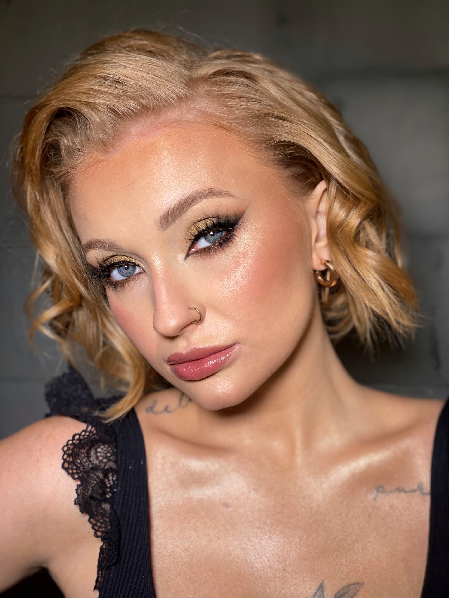 Portret kobiety z blond włosami, złotym cieniem na powiekach, czarnym eyelinerem, sztucznymi rzęsami, kolczykiem w nosie i ustami w kolorze nude. Widoczny tatuaż na dekolcie.