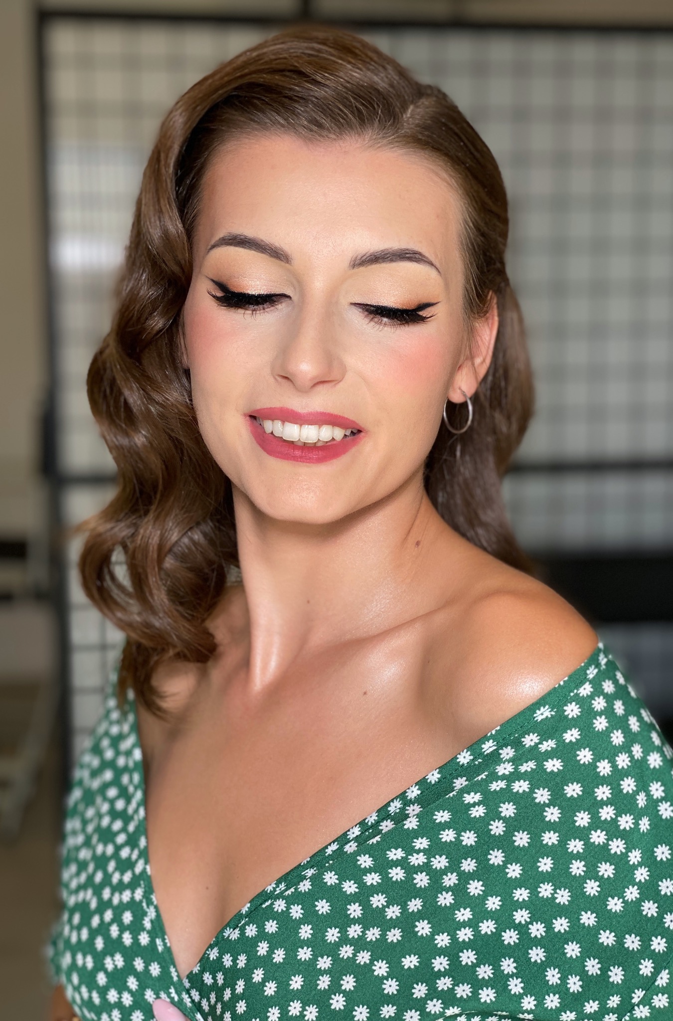 Portret uśmiechniętej kobiety z zamkniętymi oczami, w makijażu retro z wyraźną kreską eyelinerem i czerwonymi ustami, włosy ułożone w fale, ubrana w zieloną sukienkę w białe kwiatki.