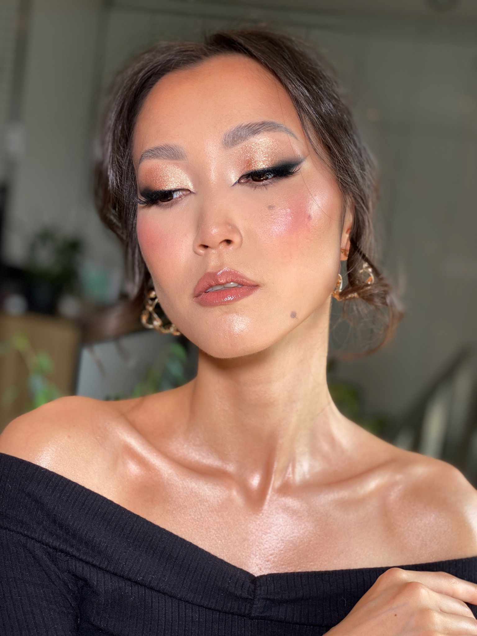 Portret kobiety z błyszczącym, złotym cieniem na powiekach, mocną czarną kreską i błyszczykiem na ustach, patrzącej w dół, z widocznymi kolczykami i odsłoniętymi ramionami.