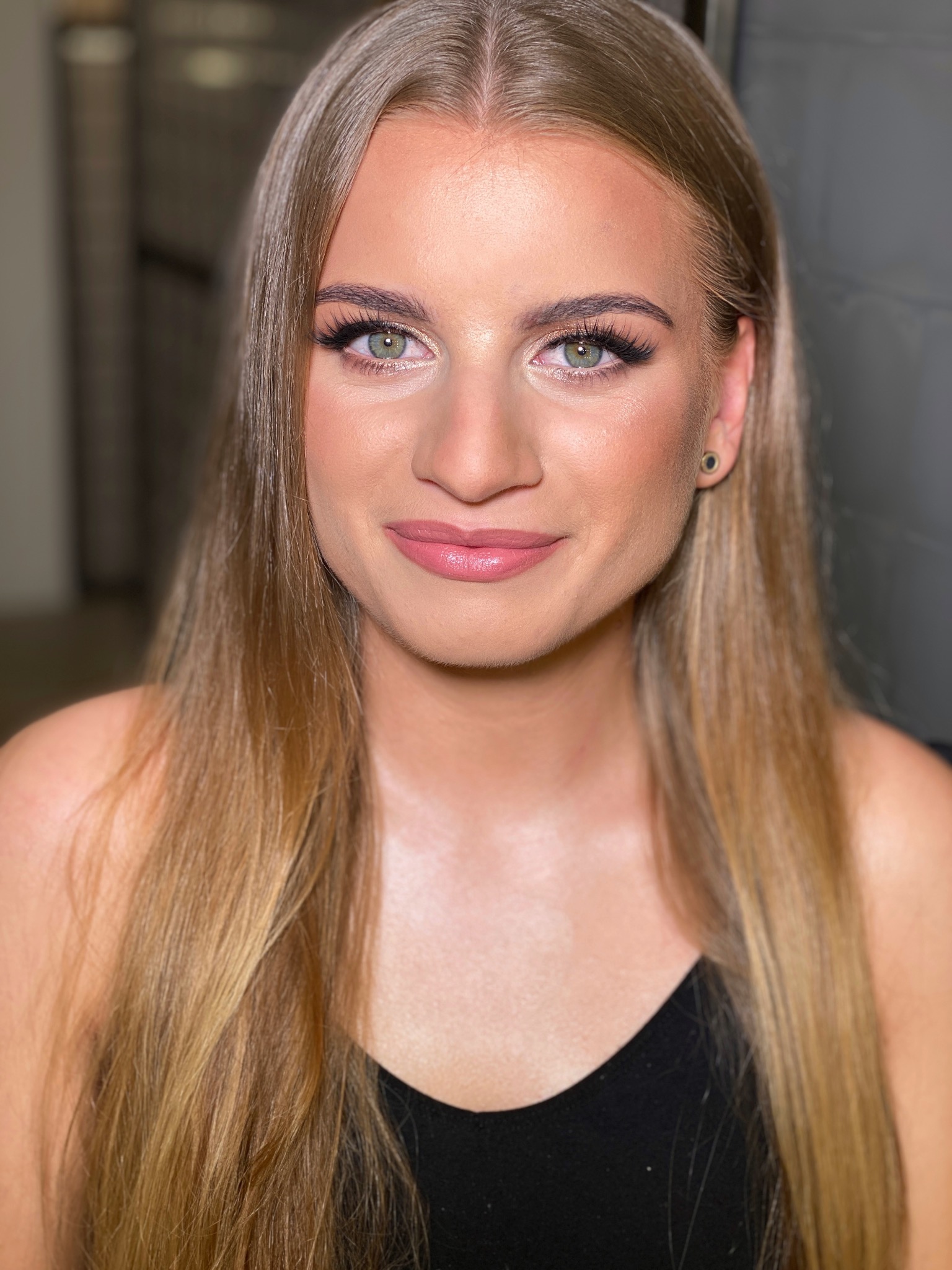 Portret młodej kobiety z delikatnym, naturalnym podkreśleniem oczu i ust, długie blond włosy, czarny top, widoczne kolczyki w uszach.