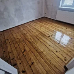 Świeżo polakierowana, błyszcząca podłoga z desek w pokoju z surowymi, szpachlowanymi ścianami i białym grzejnikiem pod oknem.