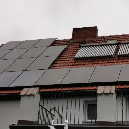 Panele fotowoltaiczne i kolektory słoneczne na dachu domu z czerwoną dachówką, widoczne z poziomu balkonu z ozdobną, metalową balustradą.