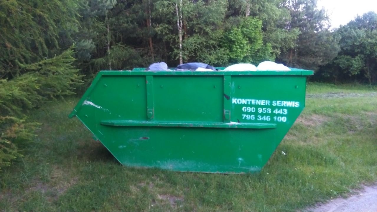 Zielony kontener napełniony odpadami stoi na trawie, z widocznym napisem 'Kontener Serwis' i numerami telefonów, w tle drzewa.