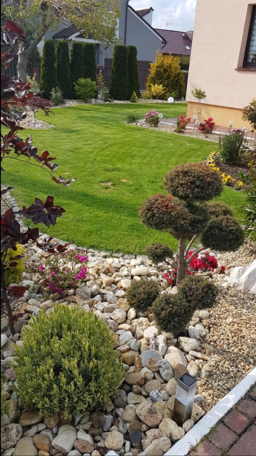 Zadbany ogród z równo przystrzyżonym trawnikiem, ozdobnymi krzewami i kamieniami. Widoczne różne gatunki roślin, lampki solarne i elementy architektury ogrodowej.