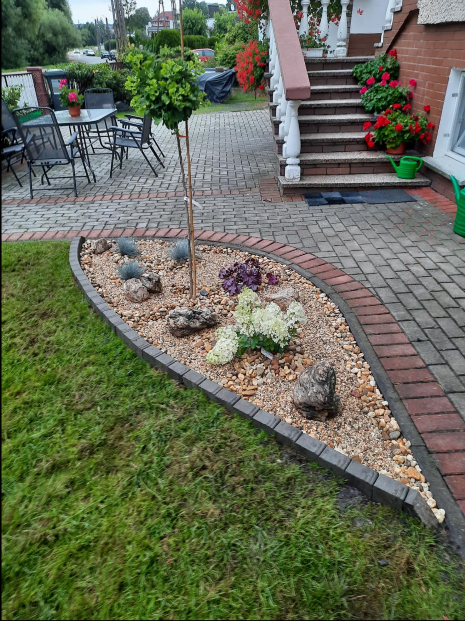 Kształtna rabata z kamykami, roślinami i ozdobnymi kamieniami, obrzeżona kostką, na tle trawnika i domu z kwiatami w donicach. Widok na ogród i otoczenie domu.