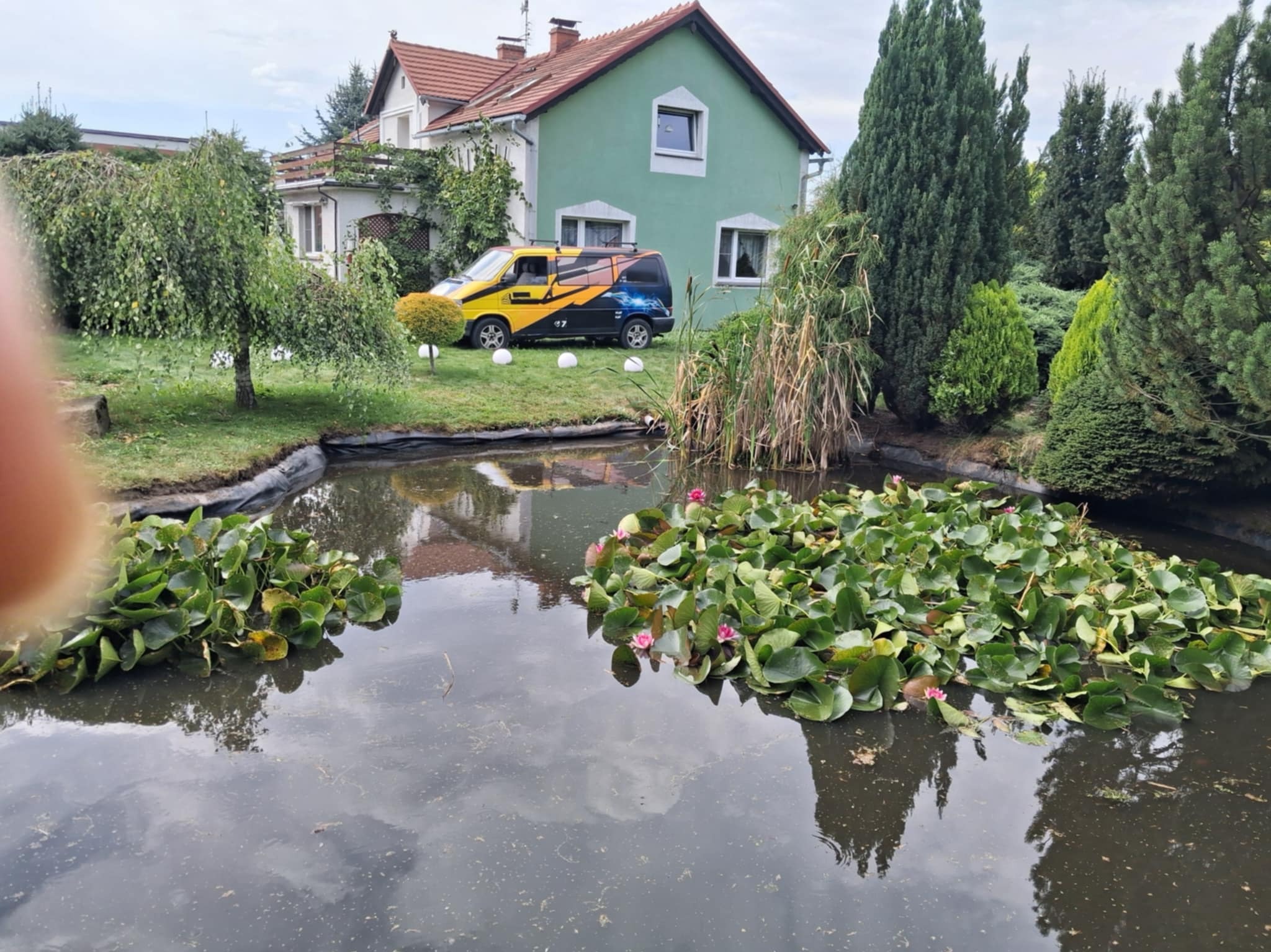 Oczko wodne z liliami wodnymi na tle domu i firmowego busa. Widoczny zadbany trawnik i różnorodne drzewa. Całość tworzy harmonijny krajobraz.