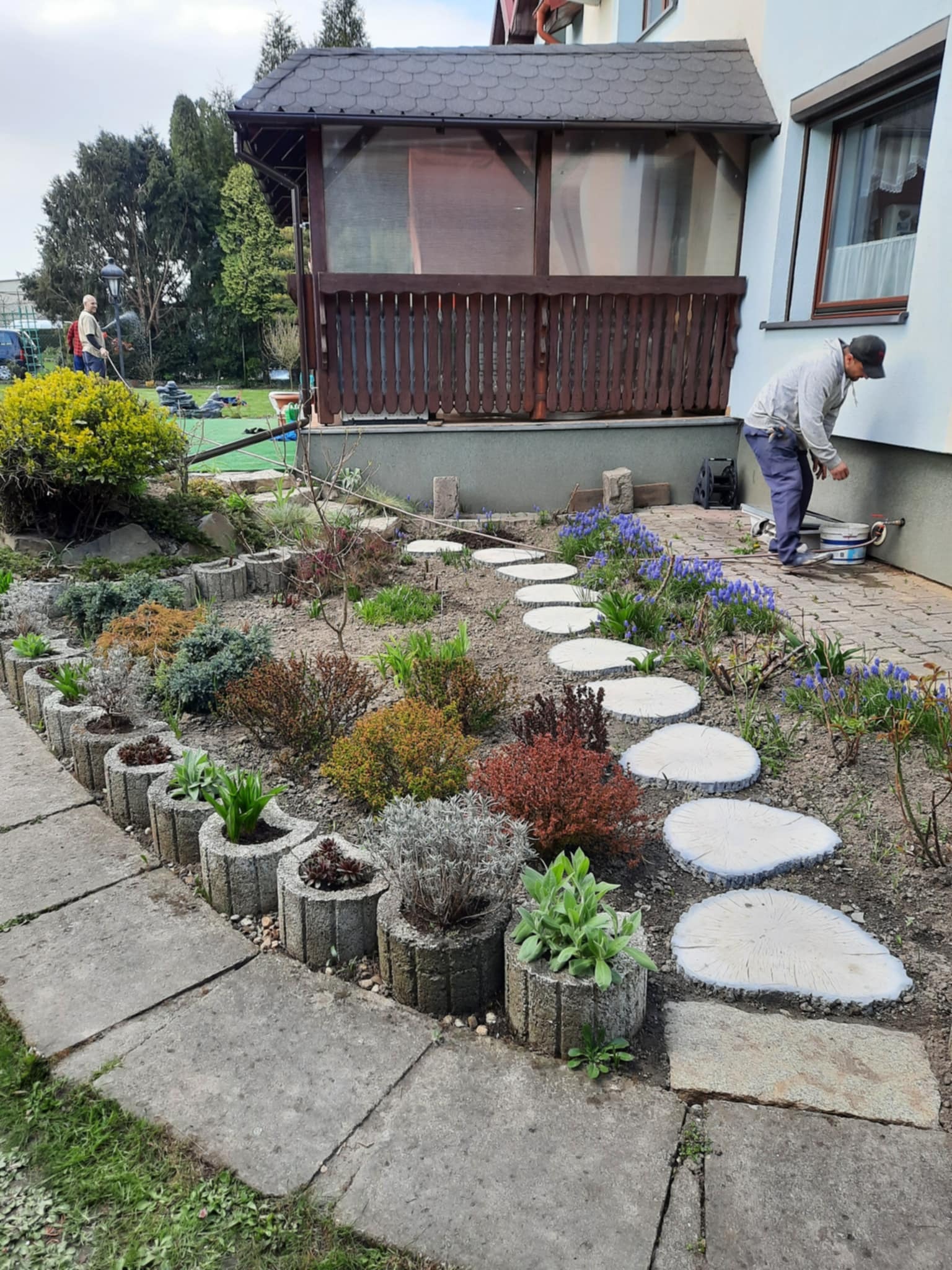 Ogród po realizacji: ścieżka z drewnianych krążków, rabaty z roślinami w betonowych donicach, budynek w tle. Widoczny mężczyzna przy ścianie budynku.