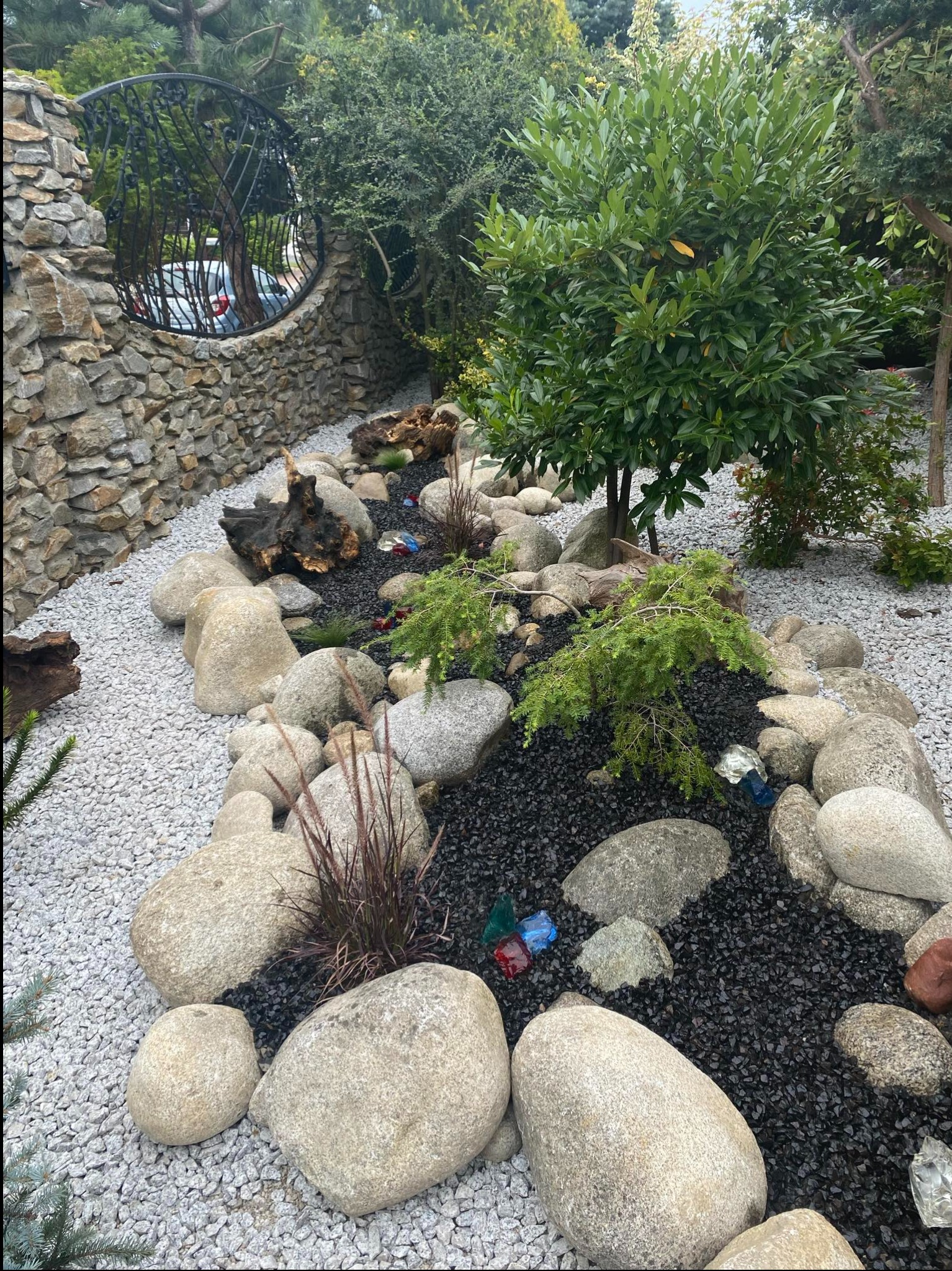 Ogród z kamienną ścieżką, ozdobnymi kamieniami, roślinami i dekoracyjnymi szklanymi elementami. W tle kamienny murek z metalową dekoracją.