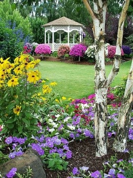 Bujny ogród z altaną w tle, kwitnące rabaty, trawnik i brzozy na pierwszym planie. Kompozycja kolorów i faktur tworzy harmonijną całość.