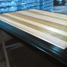 Drewniany blat z jasnych, klejonych listew, w trakcie obróbki, z czarnym, błyszczącym obramowaniem, ustawiony na metalowym stojaku w warsztacie, w tle stos zapakowanych elementów.