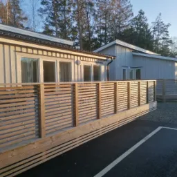 Nowoczesny dom szkieletowy z drewnianym tarasem i ażurową balustradą, widok z parkingu w słoneczny dzień.