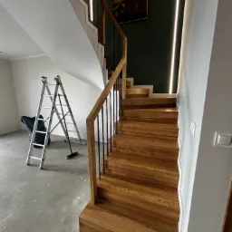 Nowoczesne, drewniane schody z czarnymi balustradami i oświetleniem LED wzdłuż ściany. W tle drabina malarska i obraz na ścianie. Widok z dołu.