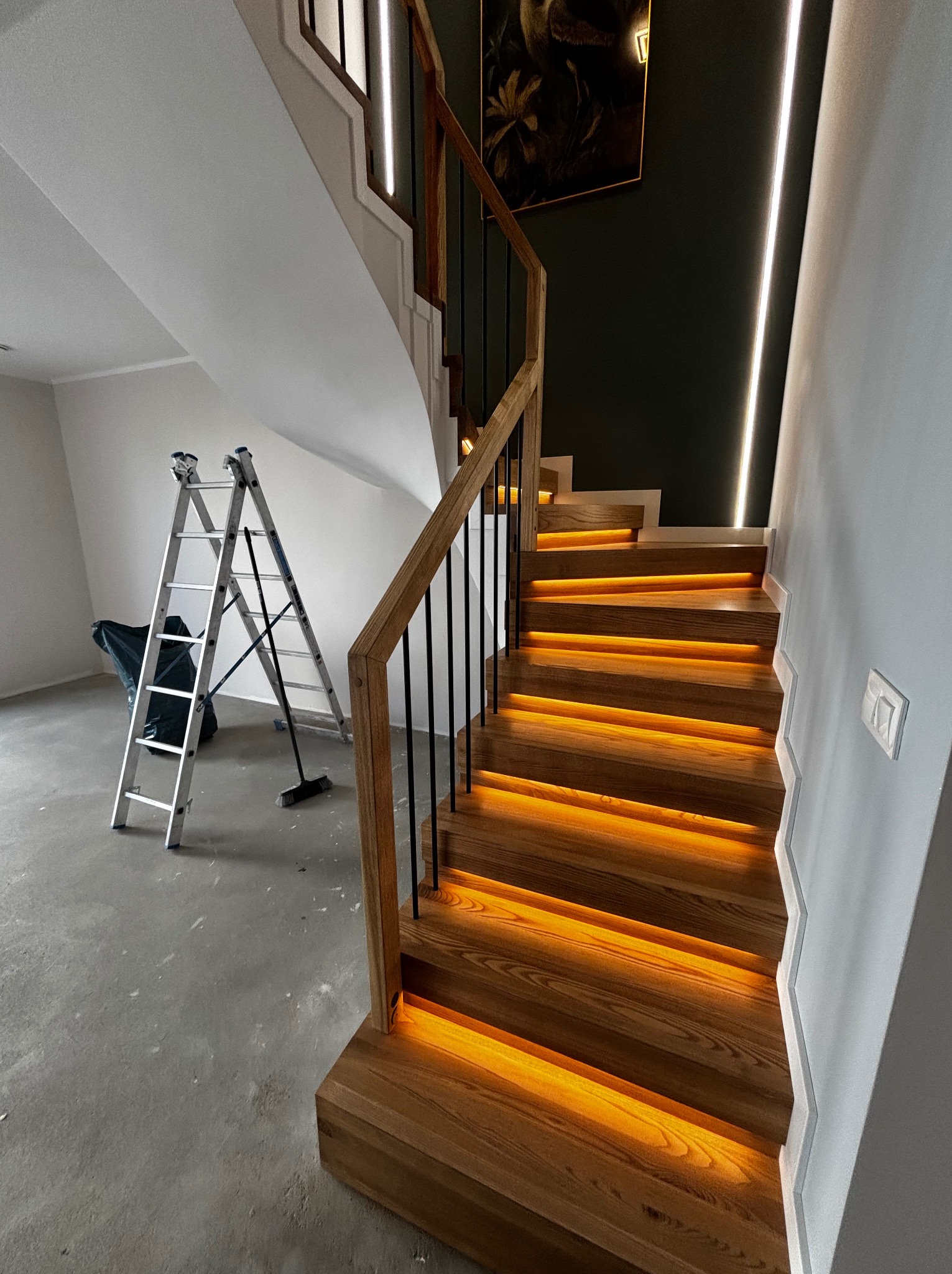 Nowoczesne schody drewniane z podświetleniem LED, czarne balustrady, drabina malarska w tle. Widok z dołu, podkreślający detale wykonania i oświetlenie.
