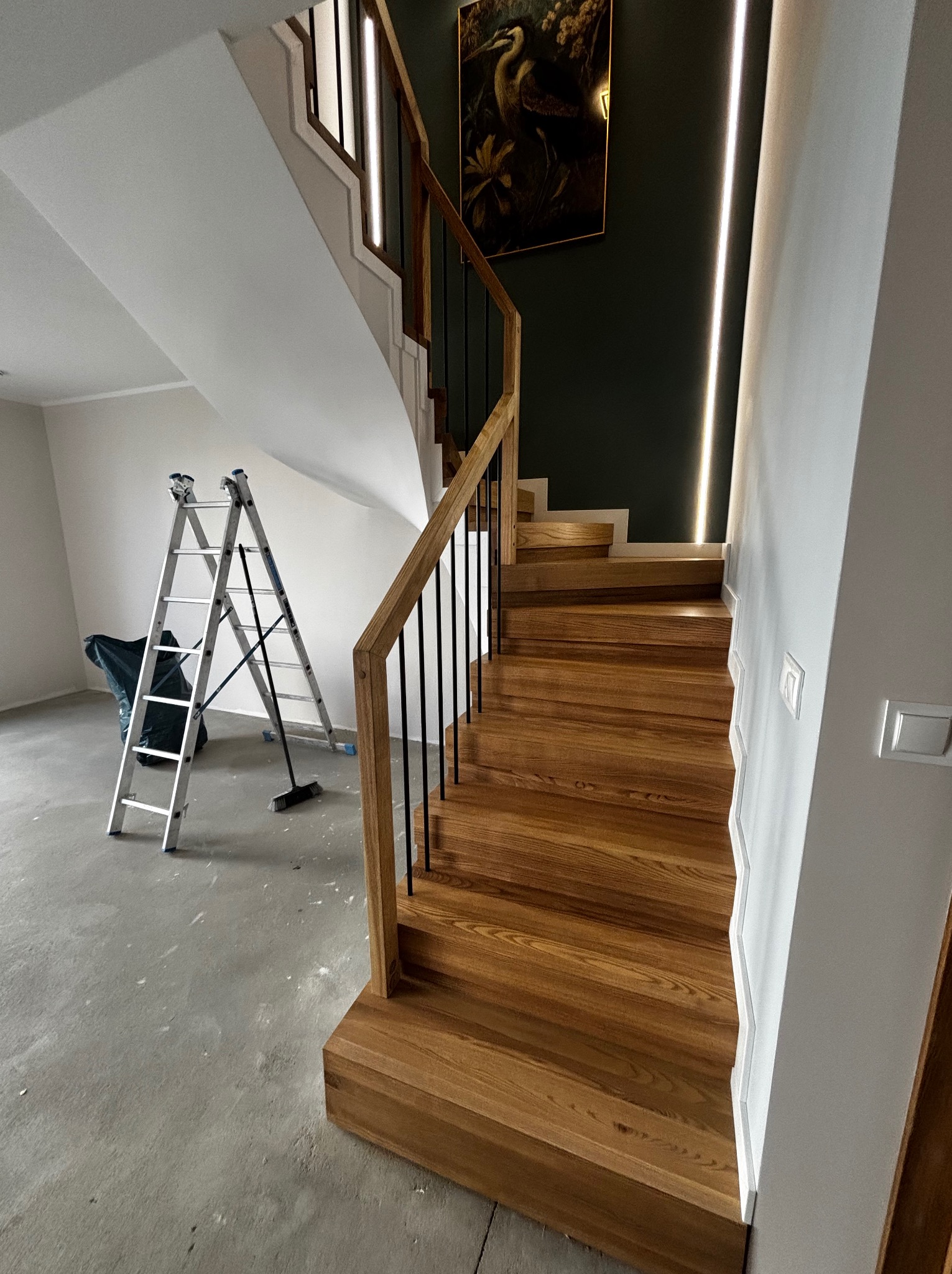 Nowoczesne, drewniane schody z czarnymi balustradami i oświetleniem LED wzdłuż ściany. W tle drabina malarska i obraz na ścianie. Widok z dołu.