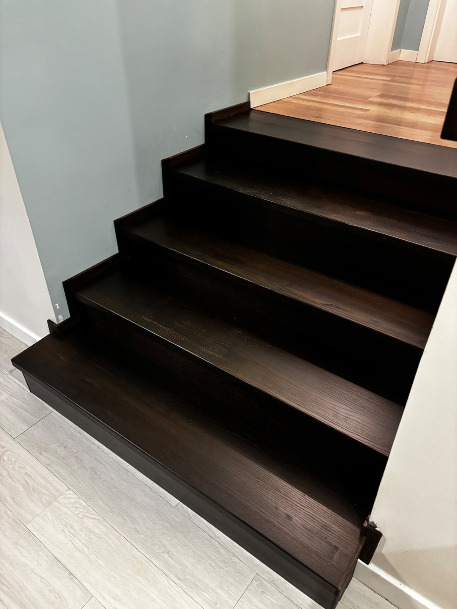 Ciemne, drewniane schody z widoczną strukturą drewna, prowadzące do góry. Ściana w kolorze gołębim i jasna podłoga dodają kontrastu.