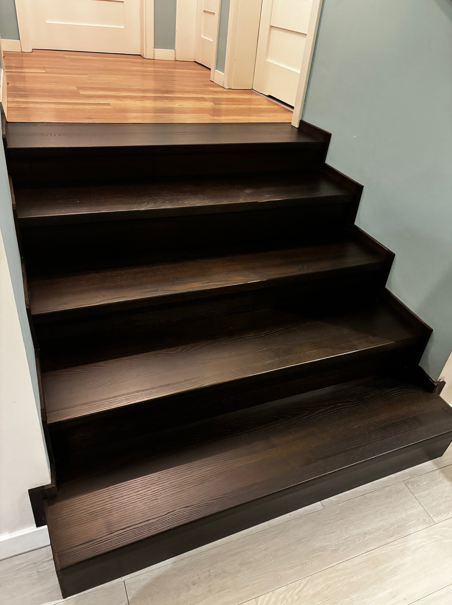 Ciemne, drewniane schody z wyraźnym usłojeniem, prowadzące do pomieszczenia z parkietem. Ściana w tle w kolorze jasnego błękitu. Ujęcie z dołu.