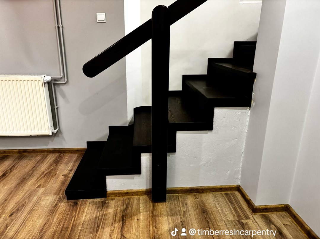 Ciemne, drewniane schody z poręczą w nowoczesnym wnętrzu. Widoczne panele podłogowe i grzejnik na szarej ścianie. Minimalistyczny design, kontrast bieli i czerni.