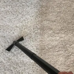 Dywan w trakcie czyszczenia: widoczna wyraźna różnica między wyczyszczoną, jaśniejszą lewą stroną a brudną, ciemniejszą prawą stroną. W kadrze fragment urządzenia czyszczącego z czarną rurą.