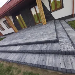 Nowoczesne schody wejściowe do domu wyłożone szarą kostką brukową z ciemnoszarym obrzeżem, prowadzące od trawnika do drzwi wejściowych.