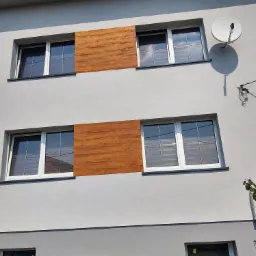 Szara elewacja domu z białymi oknami i drewnopodobnymi panelami dekoracyjnymi między nimi oraz anteną satelitarną na ścianie.