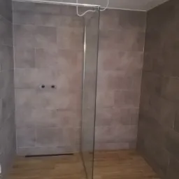 Nowoczesna kabina prysznicowa typu walk-in z szarobrązowymi płytkami na ścianach i podłodze imitującą drewno, widoczne niedokończone podłączenie oświetlenia.