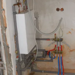 Nowa instalacja centralnego ogrzewania z rozdzielaczem podłogówki i piecem w surowym stanie, widoczne rury miedziane, zawory, oraz przewody w kolorach niebieskim i czerwonym.