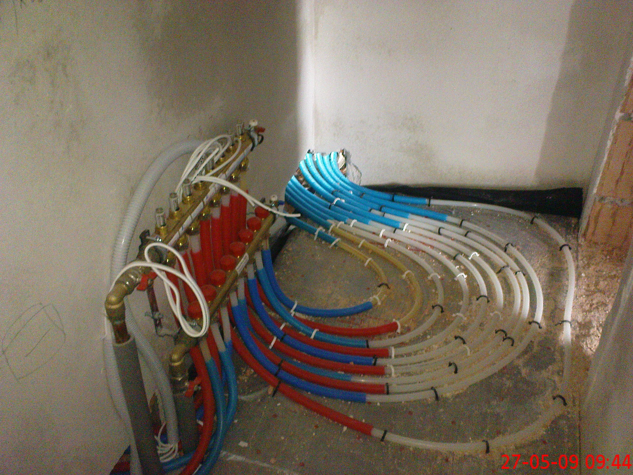 Rozdzielacz ogrzewania podłogowego z rurami PEX w kolorach czerwonym, niebieskim i białym, ułożonymi w pętle na surowej posadzce, przed wylaniem wylewki.