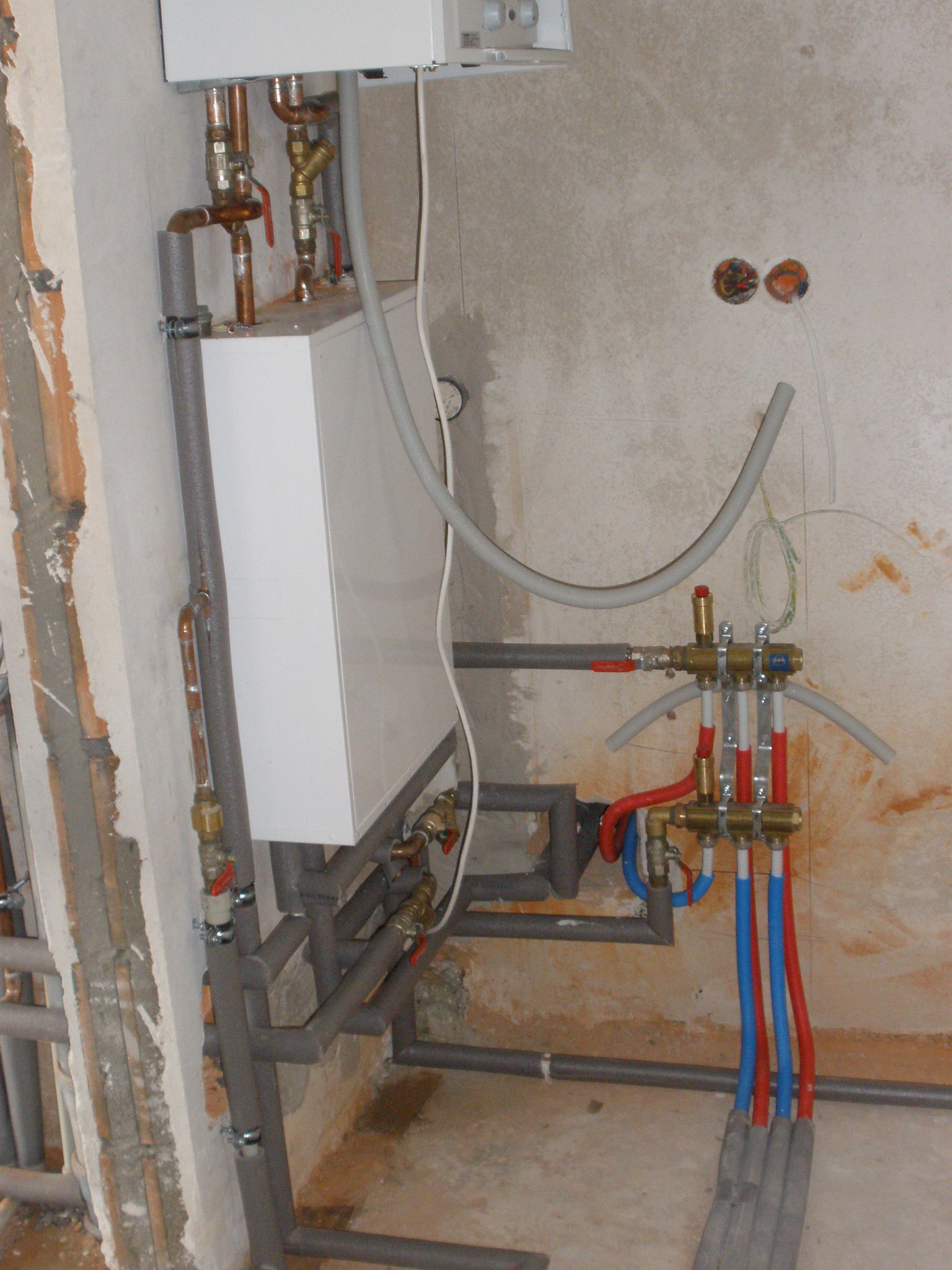 Nowa instalacja centralnego ogrzewania z rozdzielaczem podłogówki i piecem w surowym stanie, widoczne rury miedziane, zawory, oraz przewody w kolorach niebieskim i czerwonym.